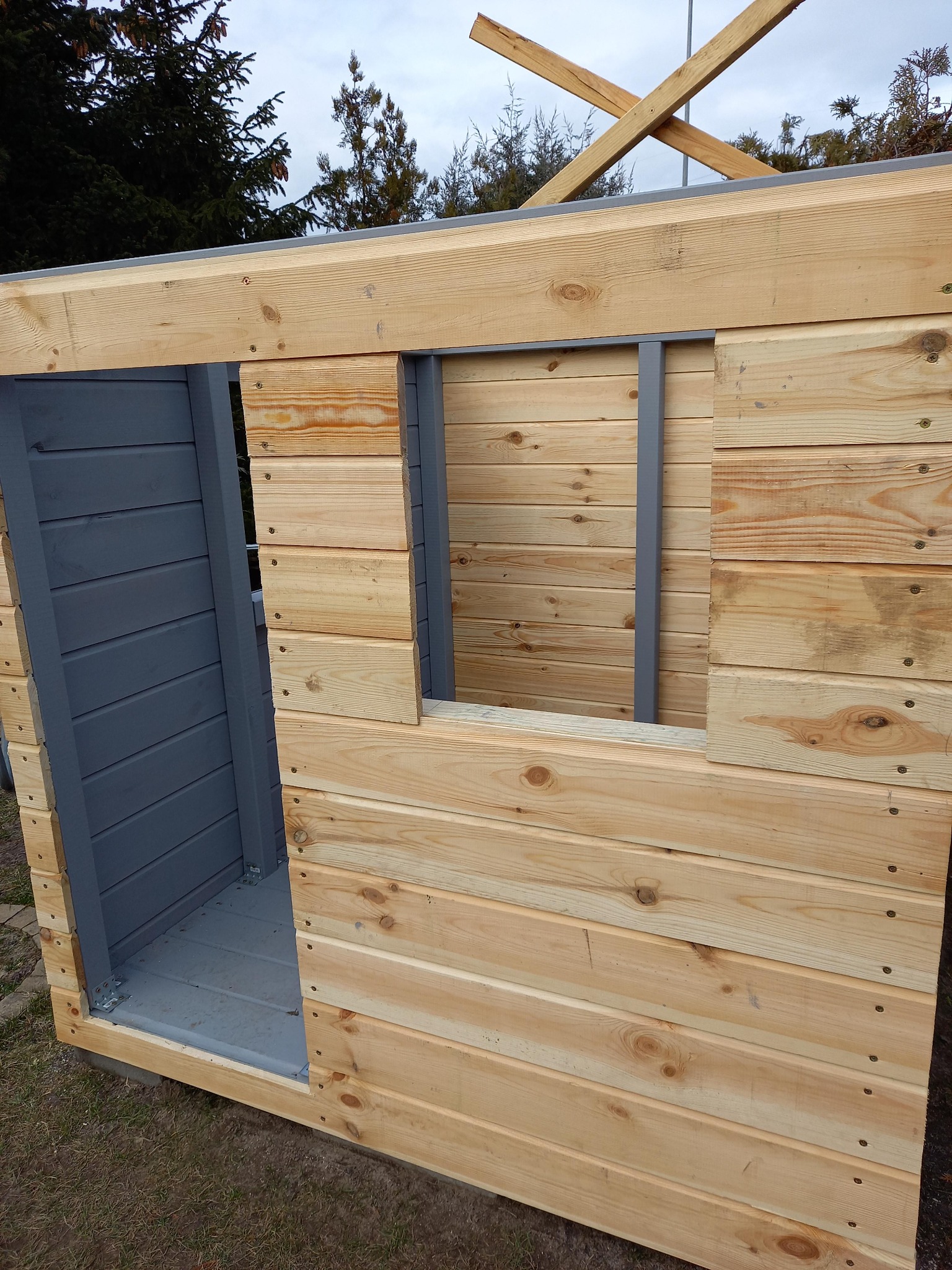 Surowa konstrukcja drewnianej altany z widocznym oknem i wejściem, częściowo pomalowana na szaro, z elementami dachu w tle. Widoczne detale łączeń i faktura drewna.