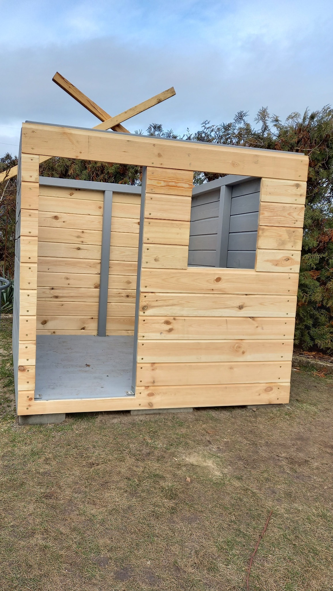 Surowa drewniana konstrukcja domku z oknem i drzwiami, z widocznymi elementami szkieletu dachu, na tle zieleni i pochmurnego nieba. Etap budowy.