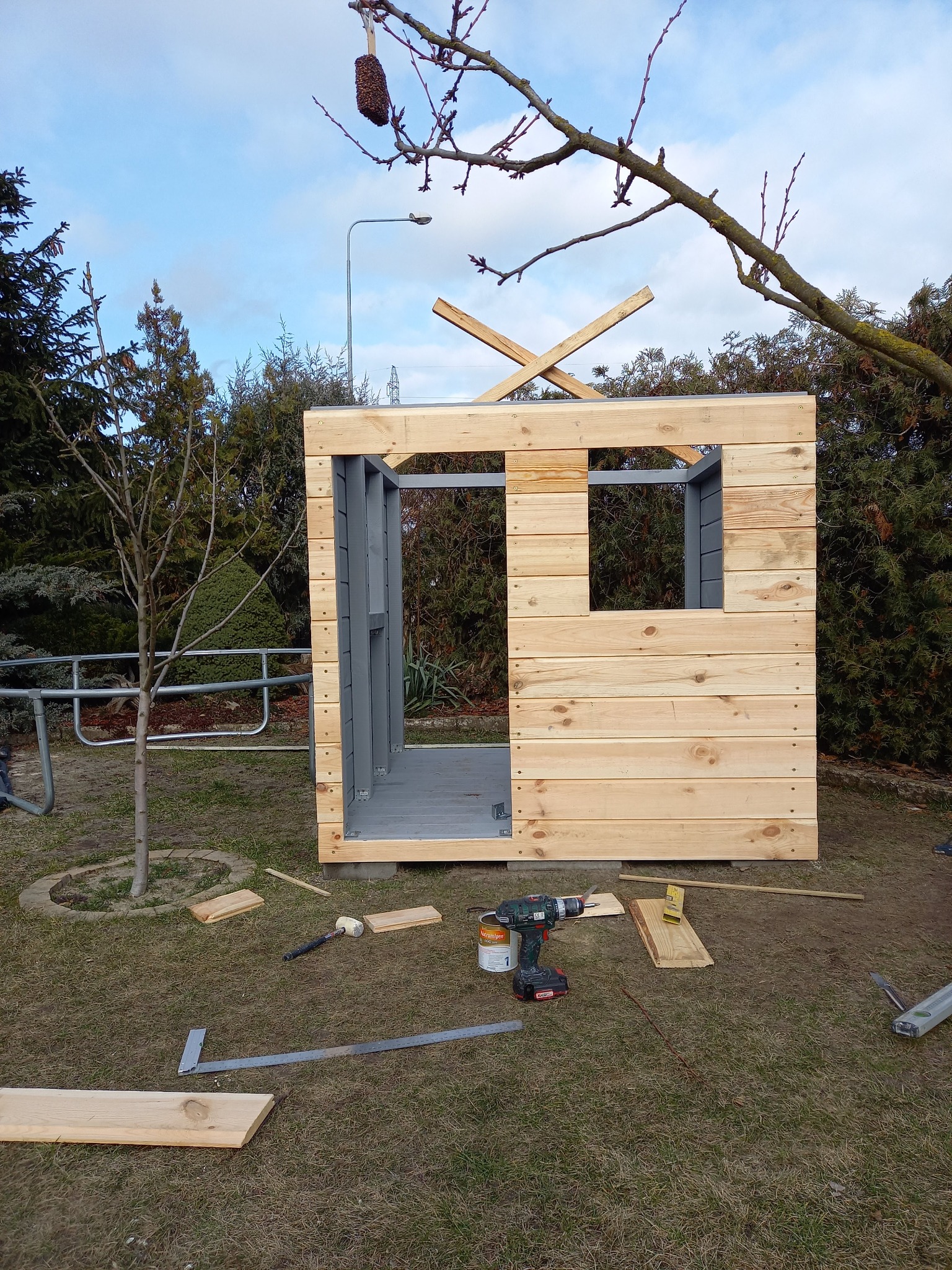 Konstrukcja altany w trakcie budowy: drewniane ściany z oknem, narzędzia na trawie, surowe drewno i szare elementy konstrukcyjne. Widok z zewnątrz, w tle drzewa.