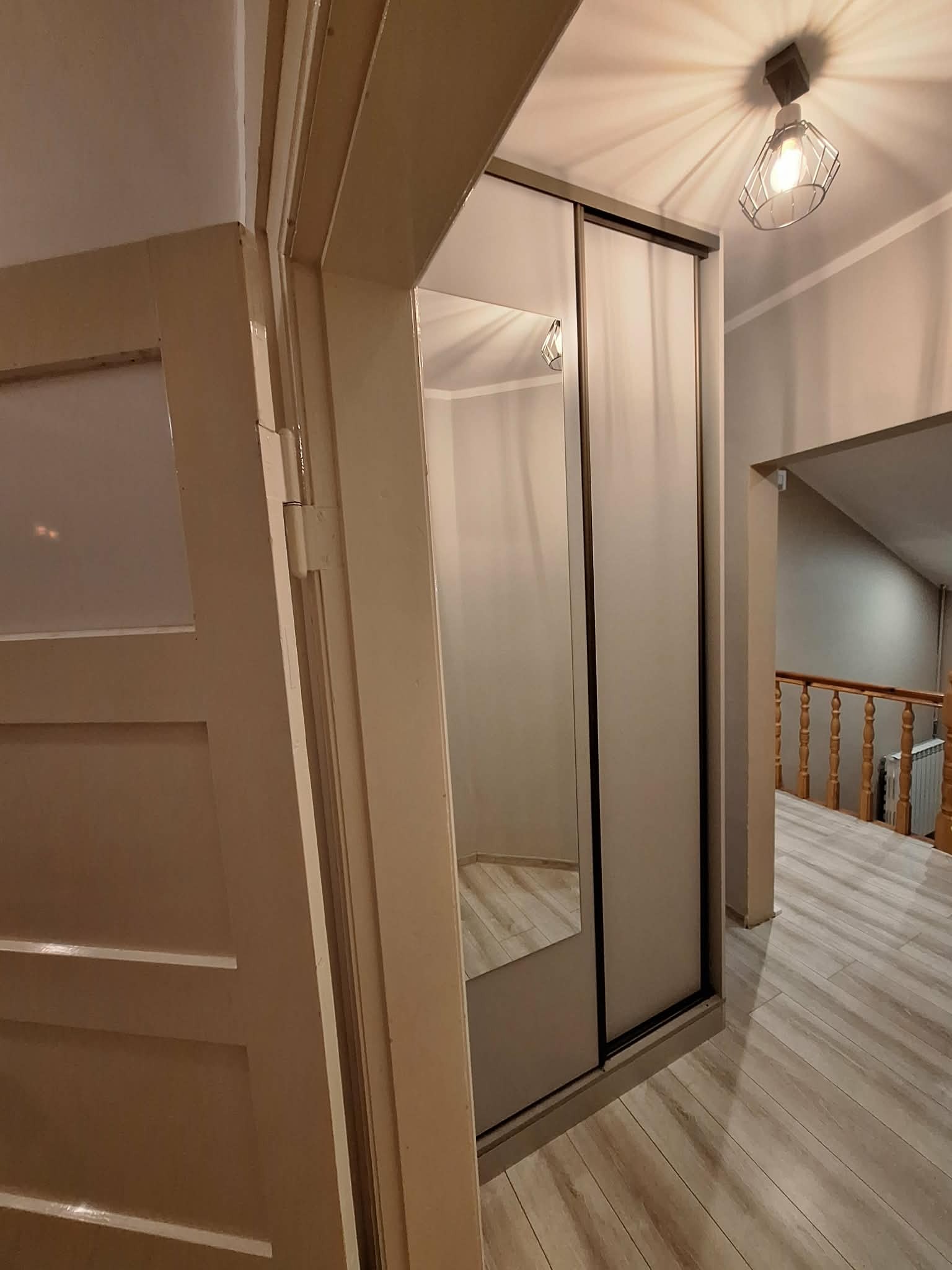 Szafa przesuwna z lustrem w przedpokoju, widok z boku otwartych drzwi. Podłoga wyłożona panelami, w tle schody z drewnianą balustradą.