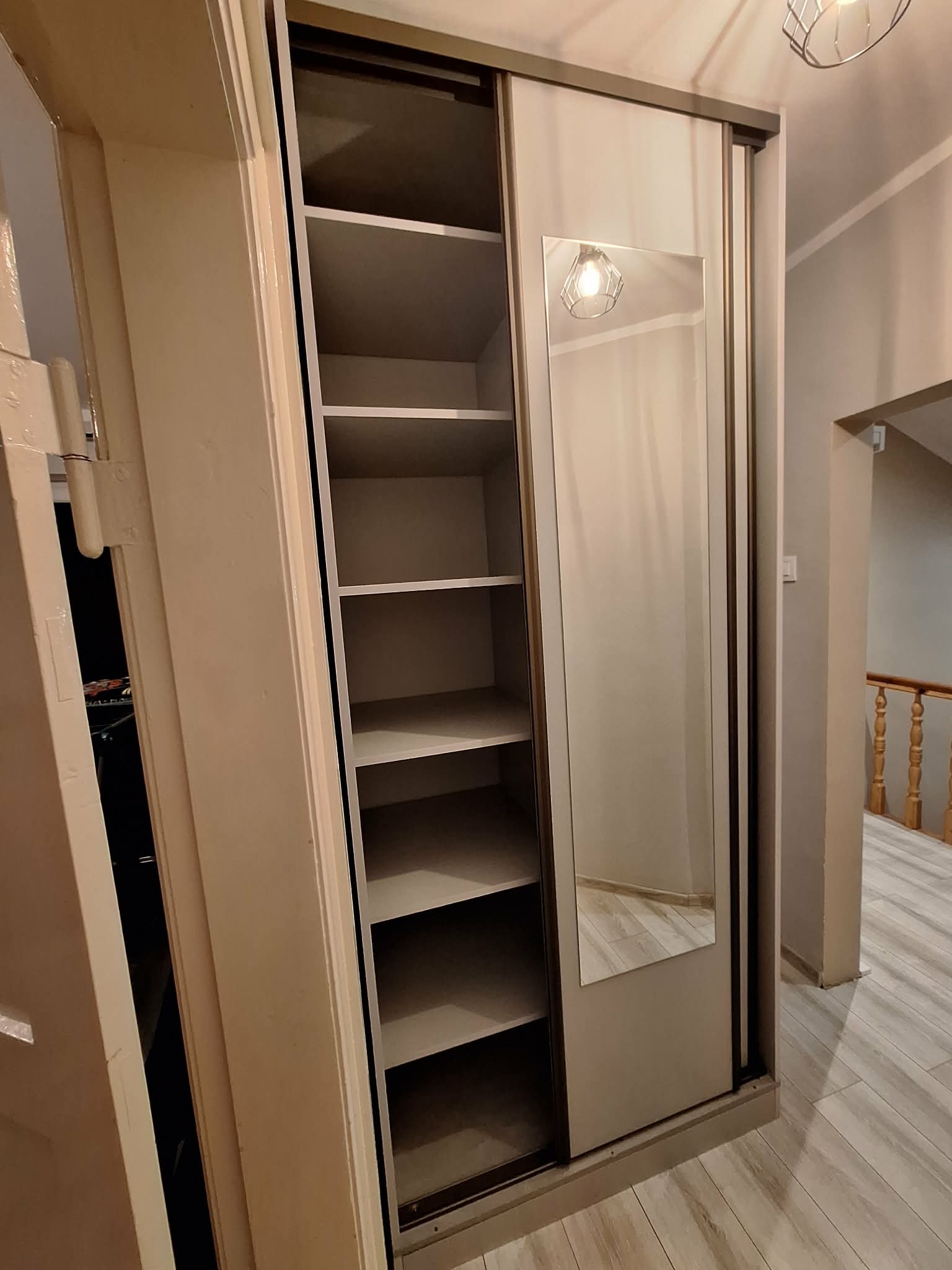 Szafa przesuwna z lustrem i otwartymi półkami w jasnym wnętrzu. Widoczny fragment podłogi i schodów. Ujęcie z bliska, prezentujące detale wykończenia.