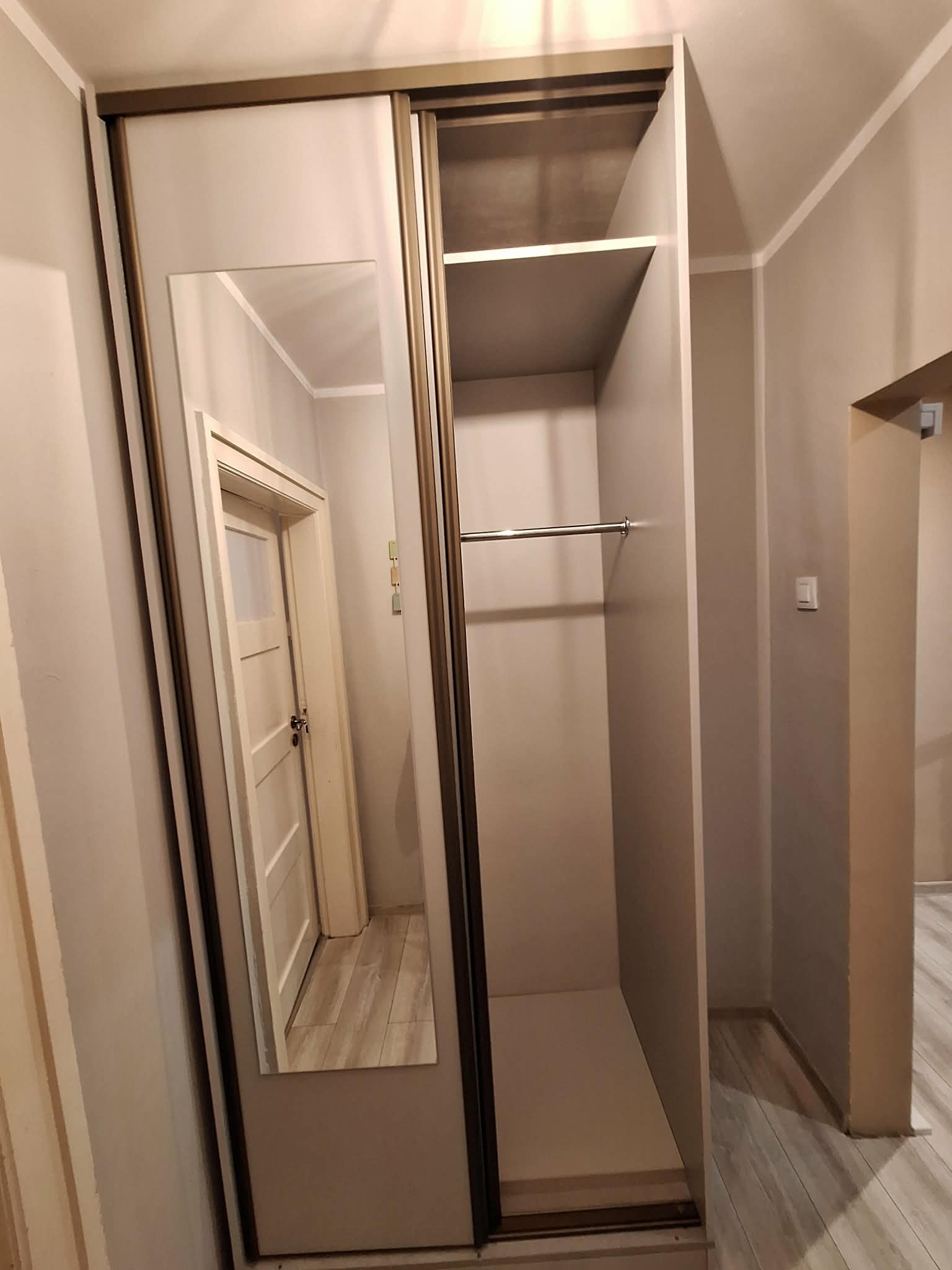 Wnętrze szafy przesuwnej z lustrem, widok na półki i drążek na ubrania. W lustrze odbija się fragment korytarza z drzwiami. Neutralna kolorystyka ścian i mebli.