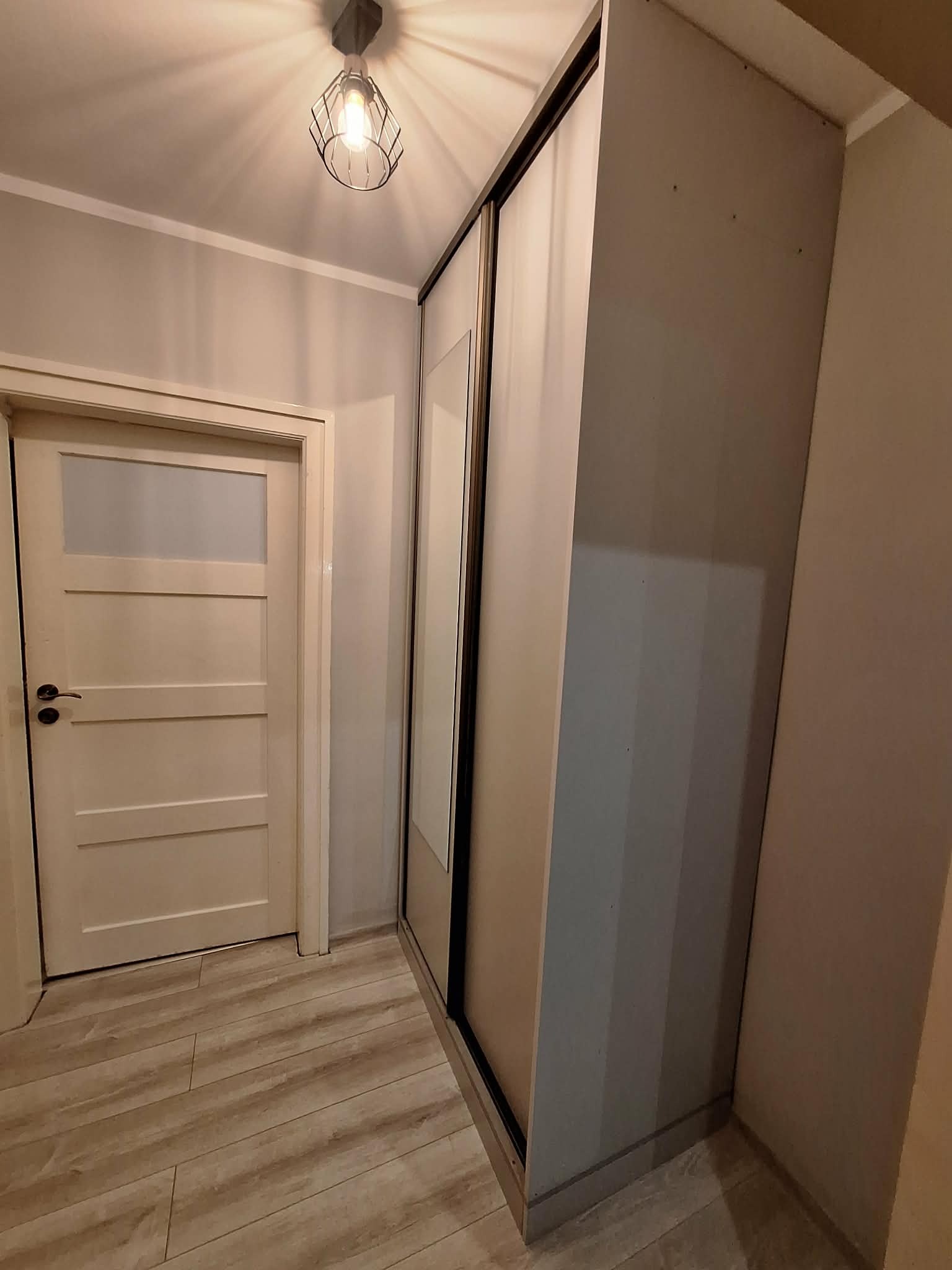 Wnętrze: zabudowa szafy przesuwnej z lustrem w przedpokoju, widoczna drewniana podłoga i drzwi. Oświetlenie sufitowe w stylu industrialnym.