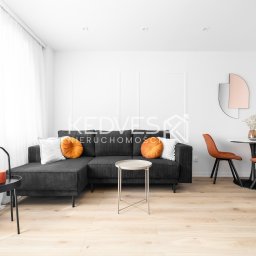 Kedves Nieruchomości Sp. z o.o. - Jasne wnętrze z szarą sofą narożną, okrągłymi pomarańczowymi poduszkami i stolikiem. W tle stół z krzesłami i dekoracyjne lustro. Minimalistyczny design z drewnianą podłogą.