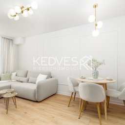 Kedves Nieruchomości Sp. z o.o. - Jasne wnętrze: sofa z stolikami kawowymi, okrągły stół z krzesłami, drewniana podłoga, białe ściany z dekoracyjnymi listwami, nowoczesne lampy. Minimalistyczny design.