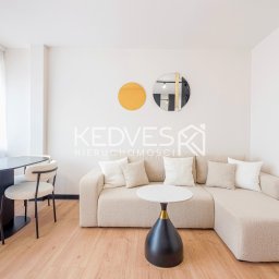 Kedves Nieruchomości Sp. z o.o. - Jasne wnętrze: beżowa sofa narożna z poduszkami, okrągły stolik kawowy, stół z krzesłami i dekoracje ścienne w postaci okręgów. Parkiet na podłodze.