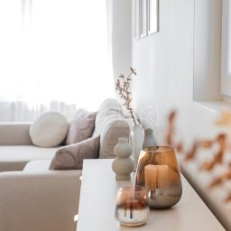 Kedves Nieruchomości Sp. z o.o. - Jasne wnętrze salonu z beżową sofą i dekoracjami: wazony, świeczniki na białej komodzie. Minimalistyczny styl, naturalne światło wpadające przez okno.