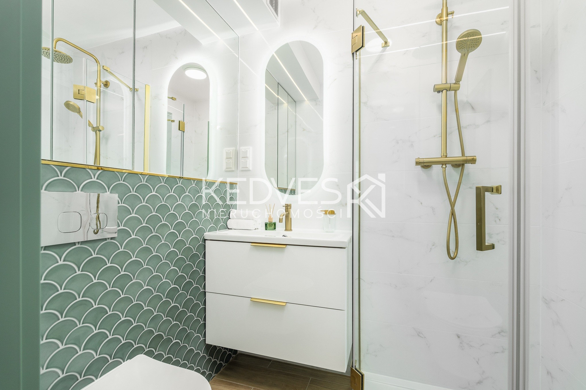 Nowoczesna łazienka z zieloną mozaiką w kształcie rybiej łuski, białymi płytkami imitującymi marmur i złotą armaturą. Oświetlone lustro nad umywalką.