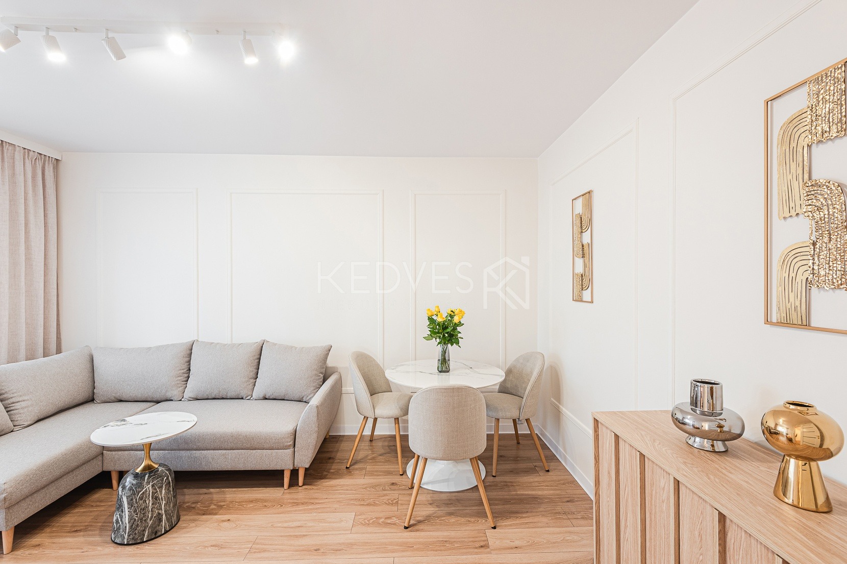 Jasne wnętrze: narożnik, stolik z marmurowym blatem, okrągły stół z kwiatami, krzesła, komoda z dekoracjami, złote obrazy na ścianie z panelami. Parkiet na podłodze.