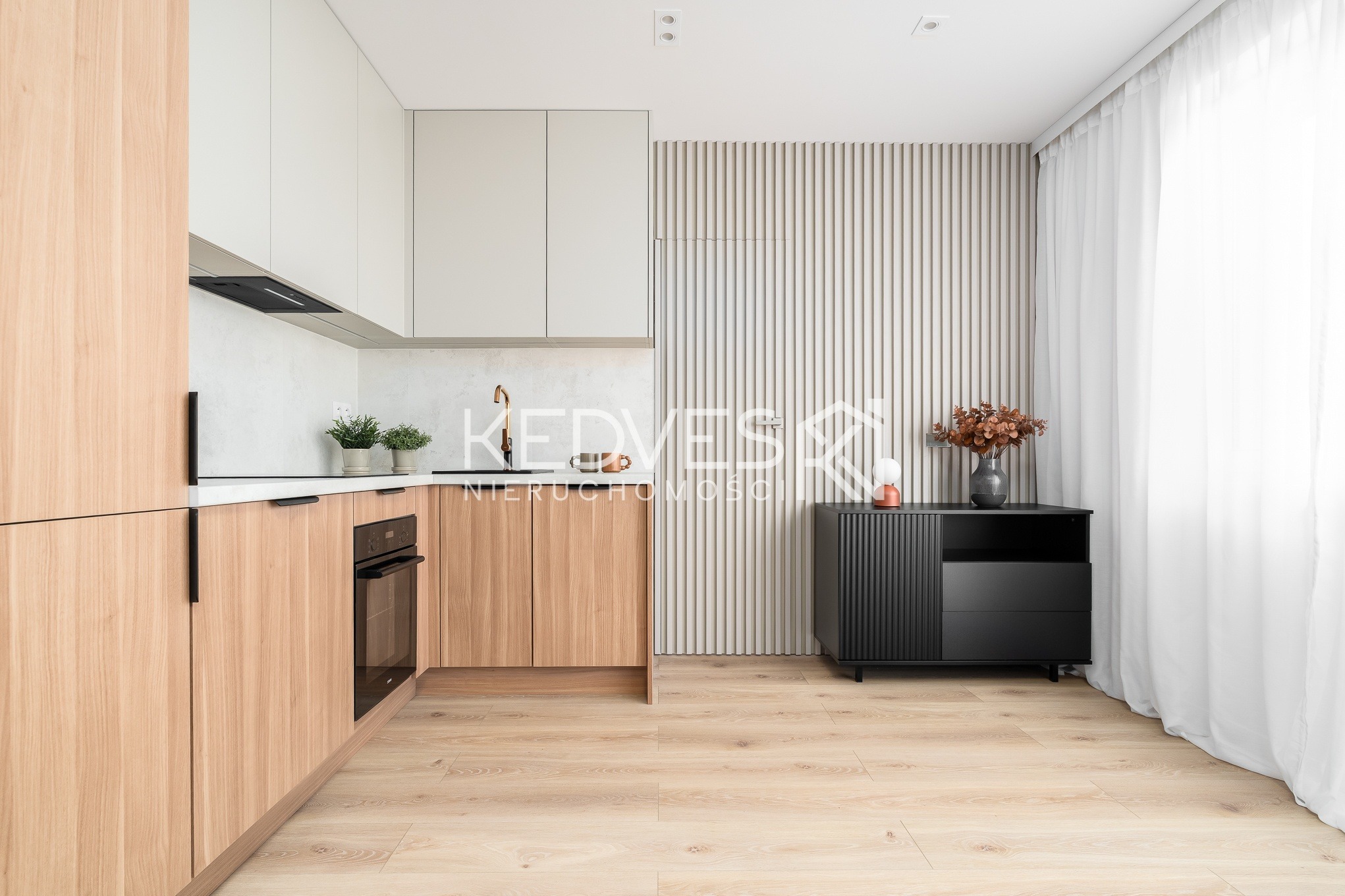 Jasna kuchnia z drewnianymi frontami szafek, białymi górnymi szafkami i czarnym kredensem na tle ściany z drewnianych listewek. Minimalistyczny design, naturalne światło.