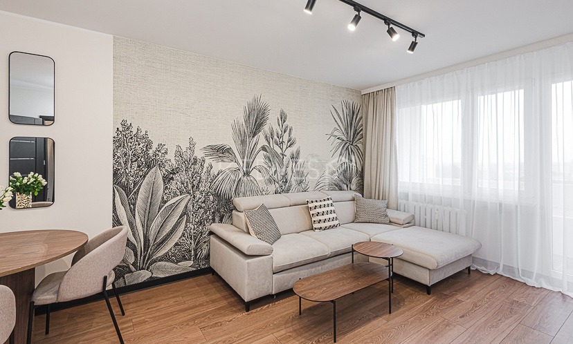 Jasny salon z drewnianą podłogą, beżową sofą narożną, stolikami kawowymi i ścianą z motywem roślinnym. Dwa lustra wiszą nad okrągłym stołem z krzesłami.