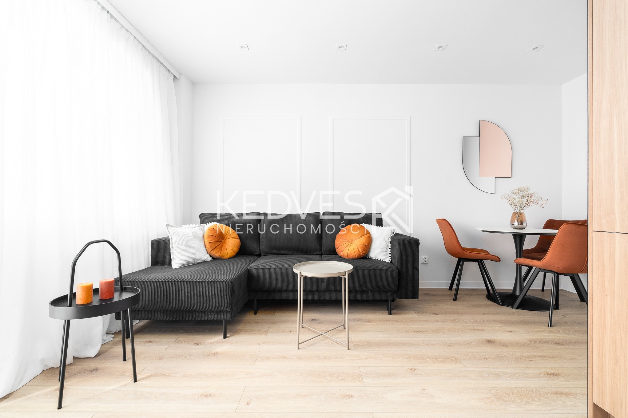 Jasne wnętrze z szarą sofą narożną, okrągłymi pomarańczowymi poduszkami i stolikiem. W tle stół z krzesłami i dekoracyjne lustro. Minimalistyczny design z drewnianą podłogą.