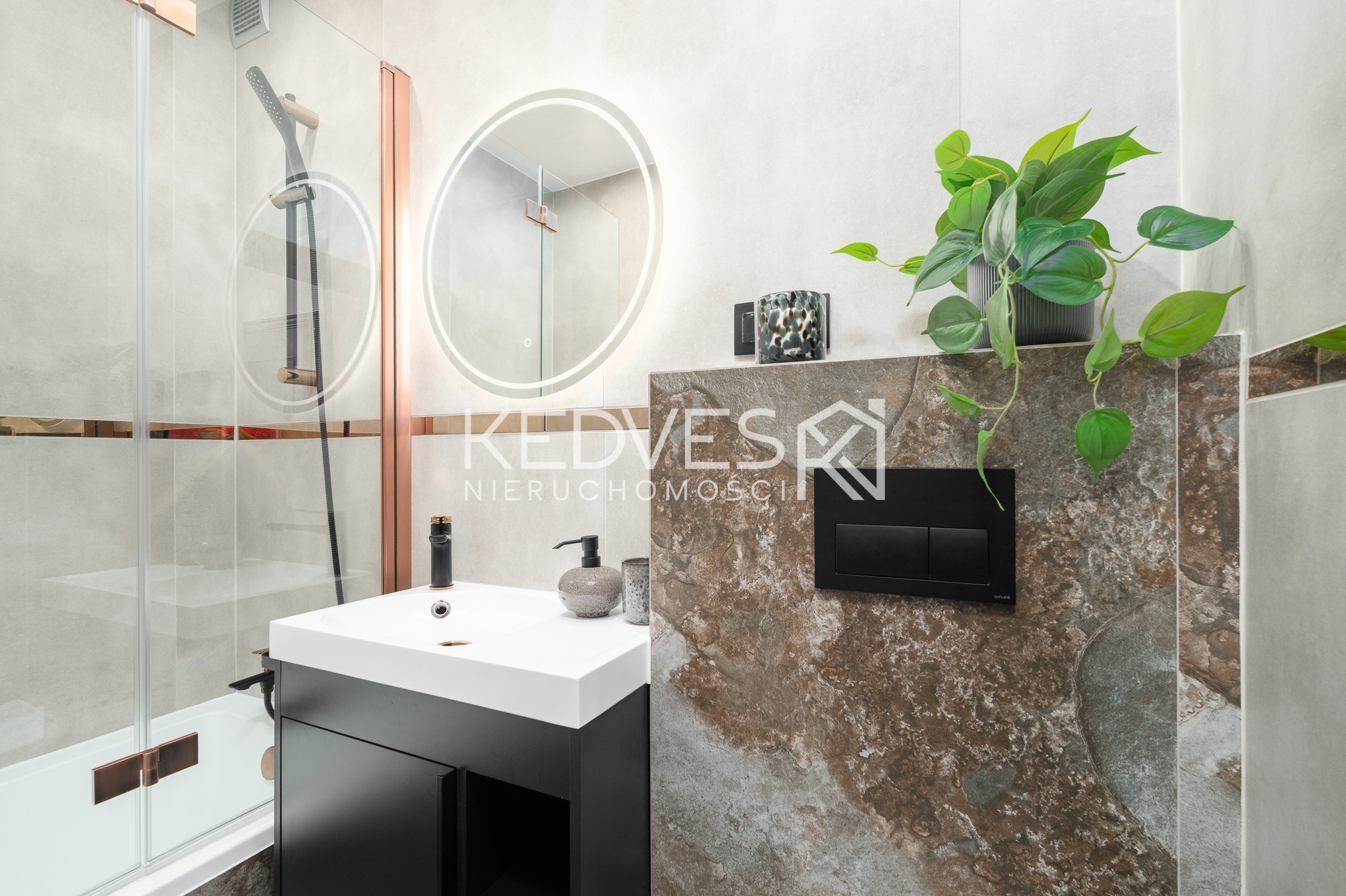Nowoczesna łazienka z okrągłym lustrem LED, czarną szafką pod umywalką i kabiną prysznicową z elementami w kolorze miedzi. Na ścianie widoczny dekoracyjny panel z rośliną.