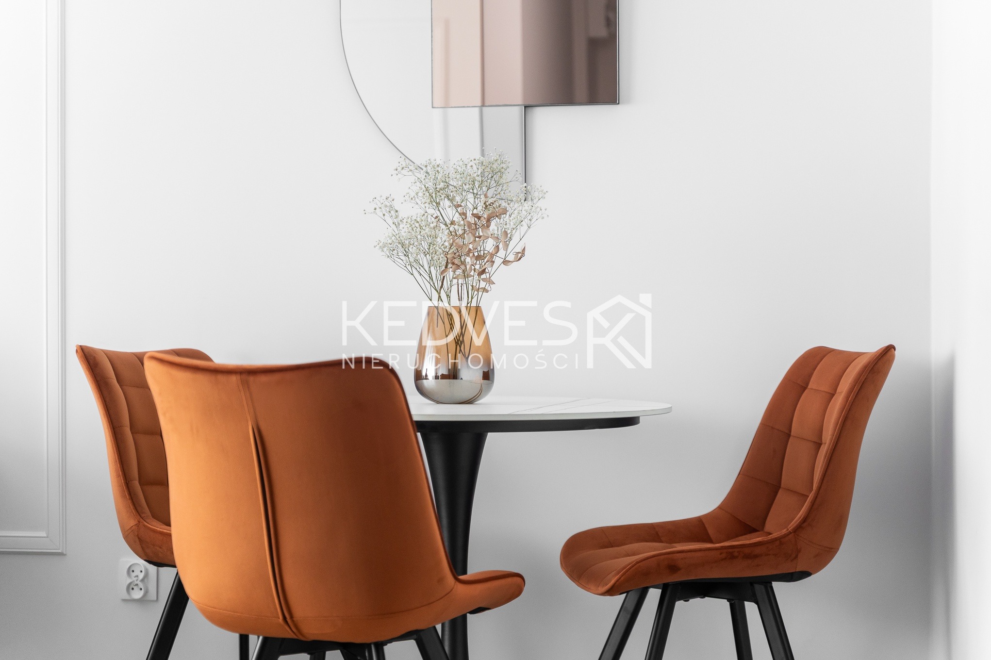 Stylowa aranżacja jadalni: okrągły stół z marmurowym blatem, bukiet gipsówki w wazonie i trzy krzesła w kolorze rdzawej pomarańczy na tle białej ściany.