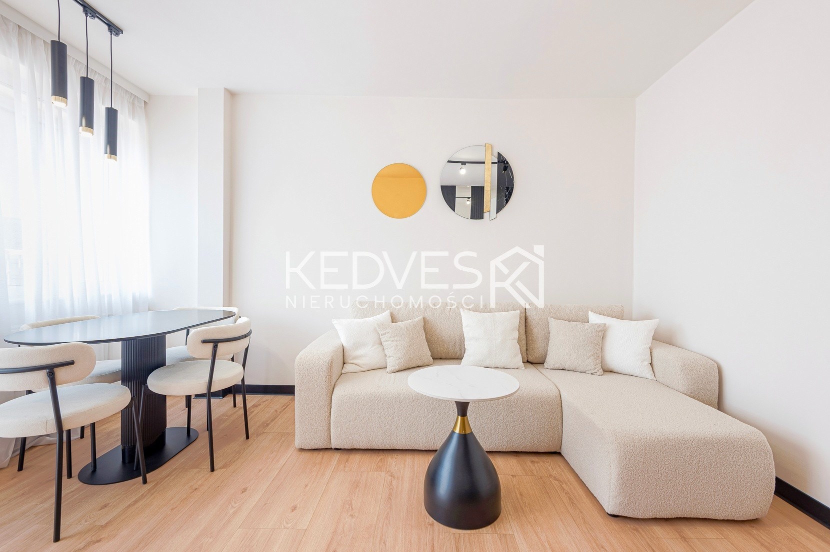 Jasne wnętrze: beżowa sofa narożna z poduszkami, okrągły stolik kawowy, stół z krzesłami i dekoracje ścienne w postaci okręgów. Parkiet na podłodze.