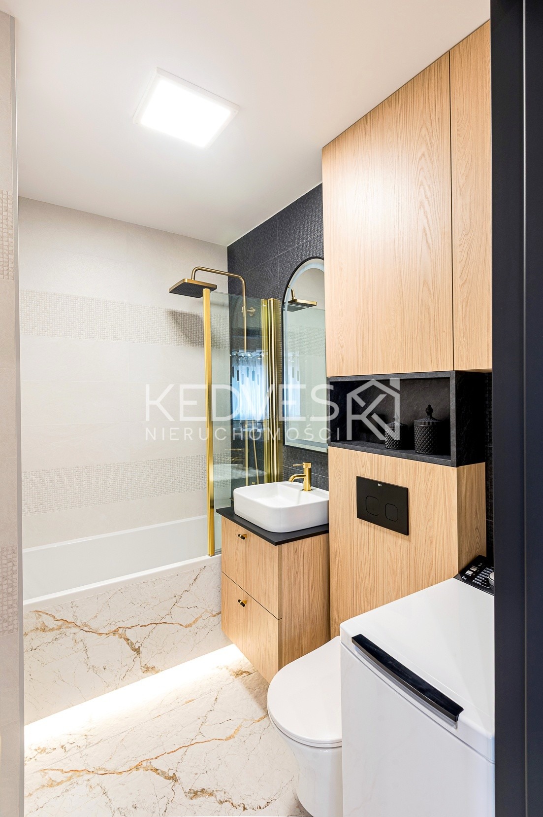 Nowoczesna łazienka z kabiną prysznicową w złotych ramach, umywalką na szafce z drewnianym fornirem i marmurową podłogą z podświetleniem LED.