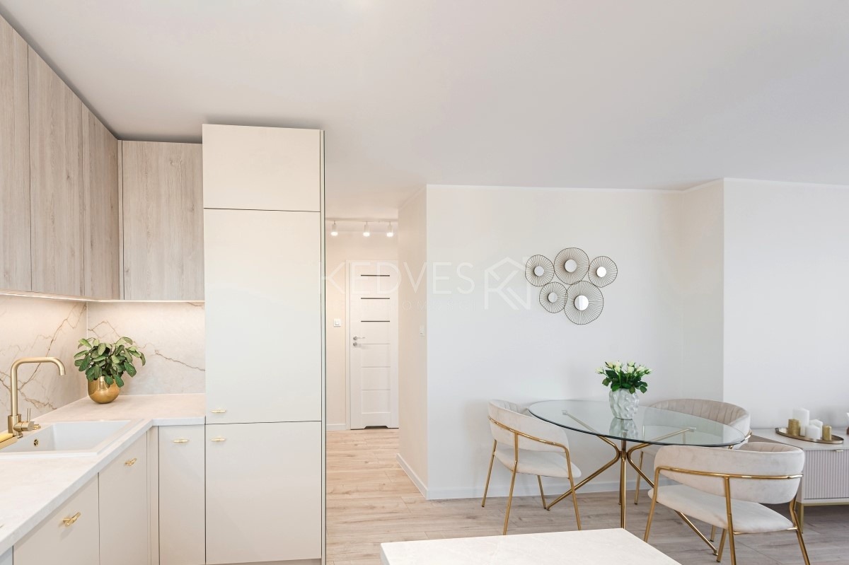 Jasna kuchnia z drewnianymi szafkami i złotymi akcentami, stół z krzesłami w stylu glamour, dekoracja ścienna z luster, minimalistyczny design.