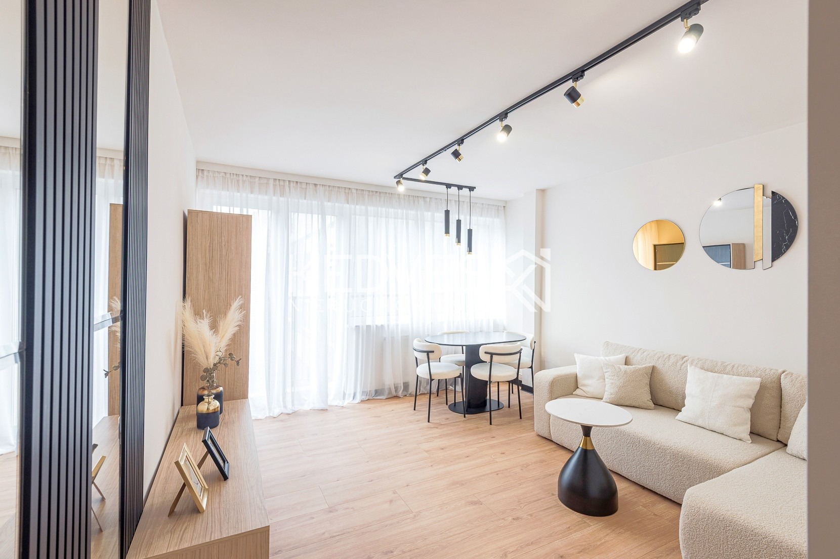 Jasne wnętrze salonu z drewnianą podłogą, beżową sofą narożną, okrągłym stołem i krzesłami. Na ścianie dekoracyjne lustra i oświetlenie szynowe.