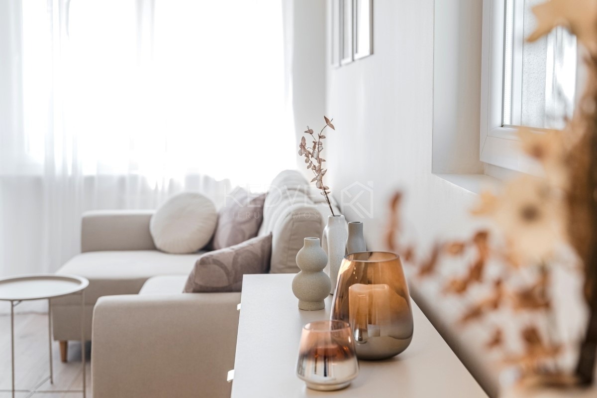 Jasne wnętrze salonu z beżową sofą i dekoracjami: wazony, świeczniki na białej komodzie. Minimalistyczny styl, naturalne światło wpadające przez okno.