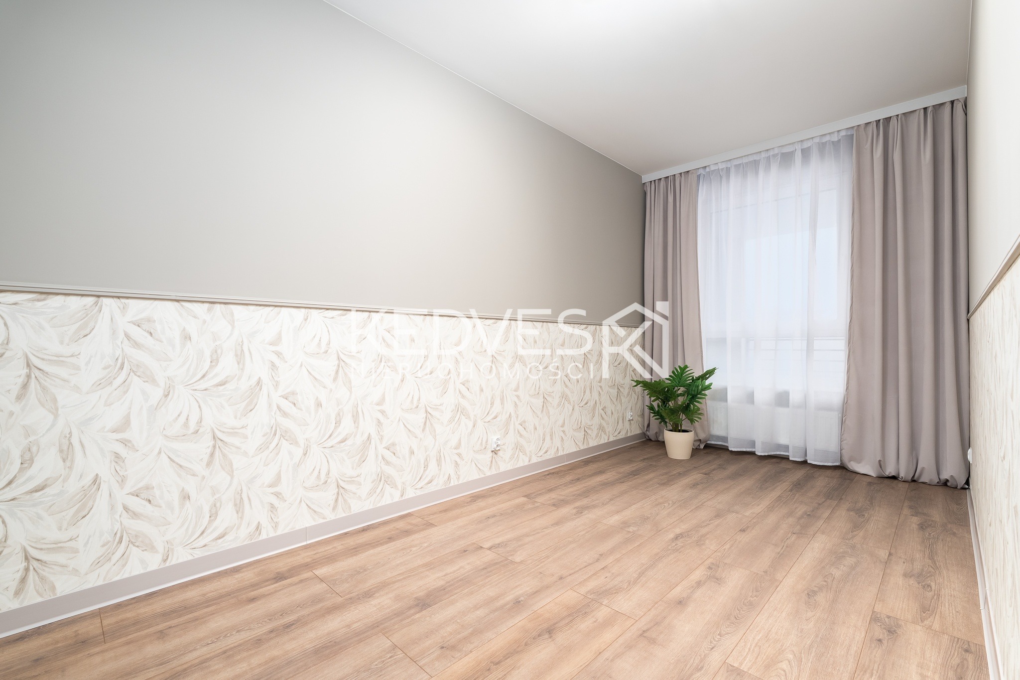 Jasne wnętrze pokoju po remoncie: panele podłogowe, tapeta w liście, szare zasłony i roślina doniczkowa przy oknie. Widok na odnowione pomieszczenie.