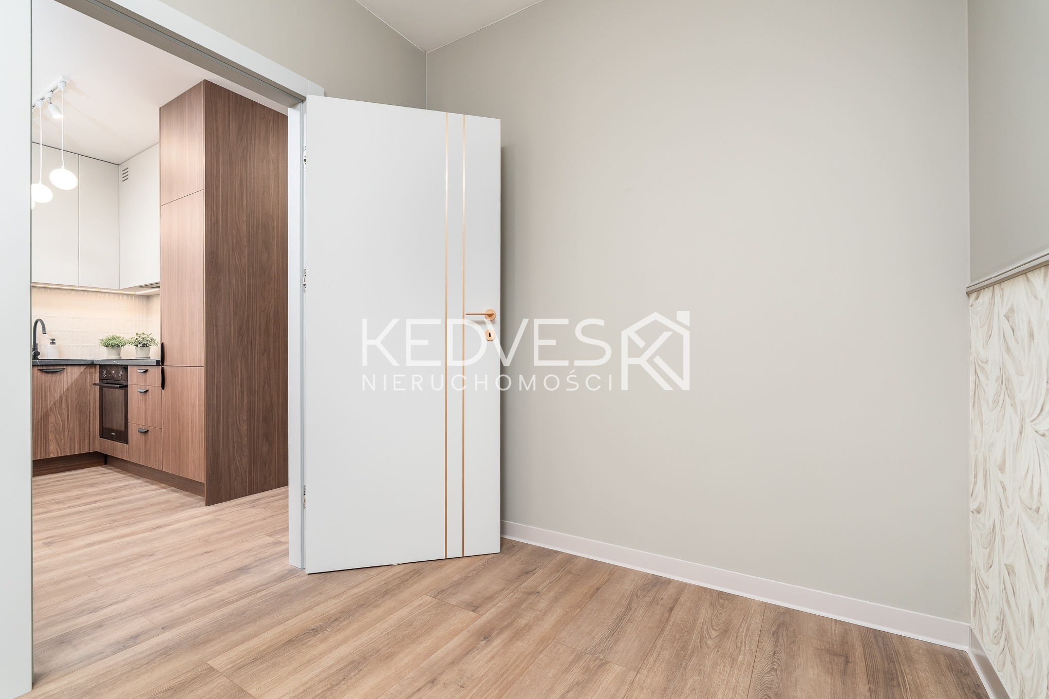 Nowoczesne wnętrze: parkiet, białe drzwi z złotymi akcentami, widok na kuchnię z drewnianymi szafkami. Minimalistyczny design z dbałością o detale.