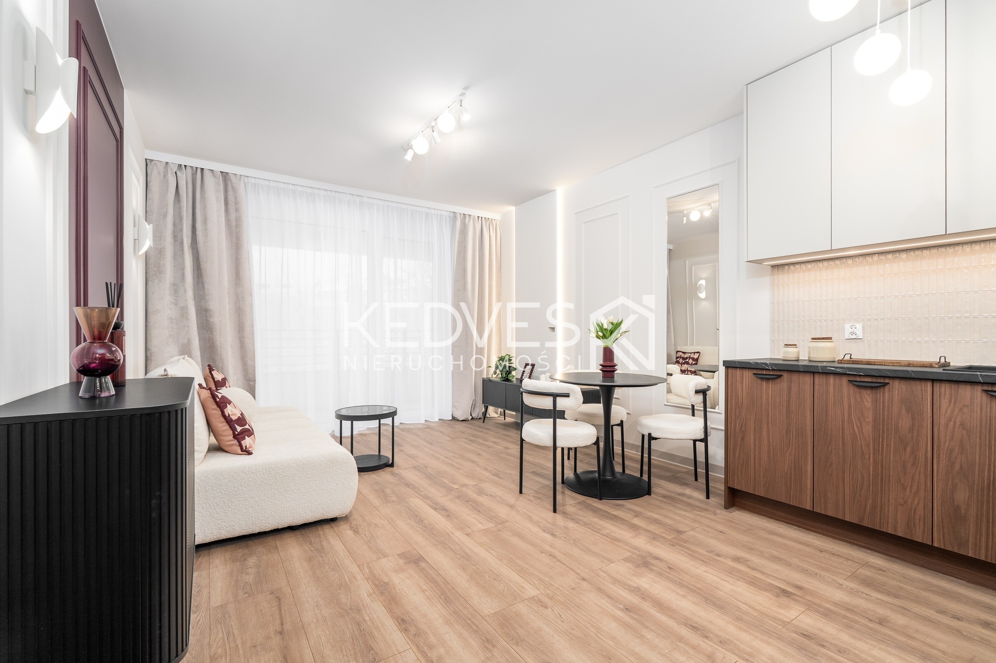 Jasne wnętrze z drewnianą podłogą: sofa boucle, okrągły stół z krzesłami, zabudowa kuchenna z drewnianymi frontami i białe ściany z dekoracyjnymi panelami.