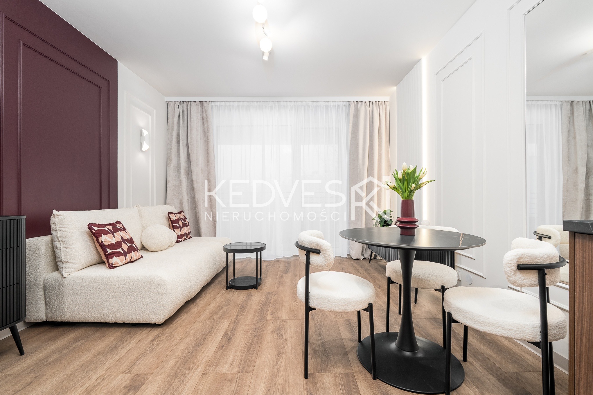 Jasne wnętrze salonu z burgundową ścianą, kremową sofą i okrągłym stołem z krzesłami w stylu bouclé. Parkiet w odcieniach brązu. Nowoczesny design.