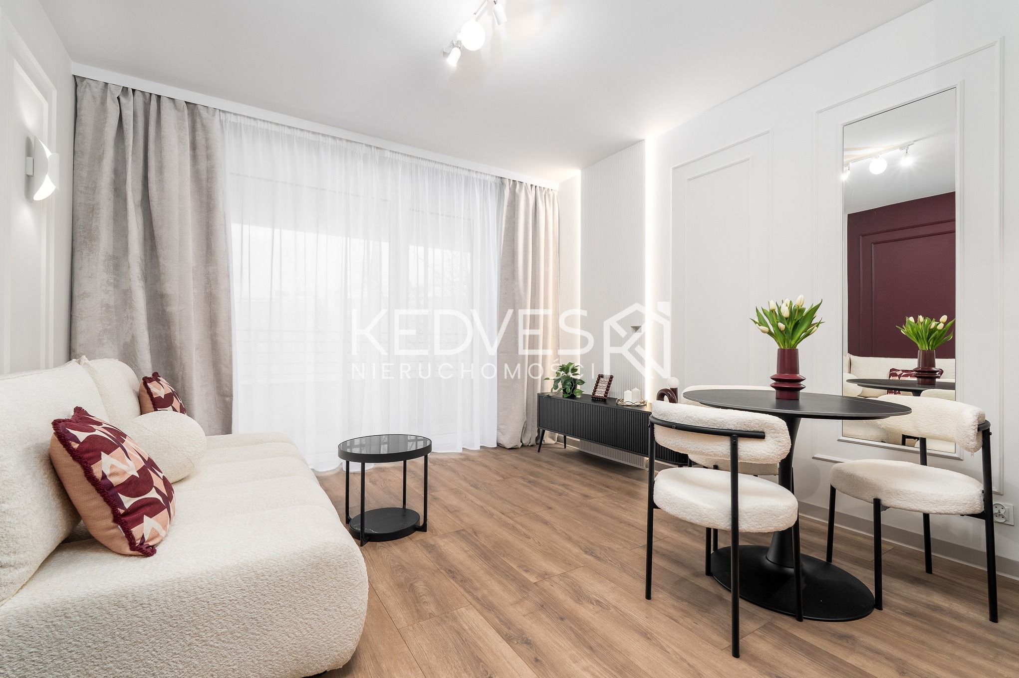 Jasne wnętrze salonu z sofą boucle, okrągłym stolikiem i czarnym stołem z kwiatami. Parkiet na podłodze, minimalistyczny design, neutralna kolorystyka.