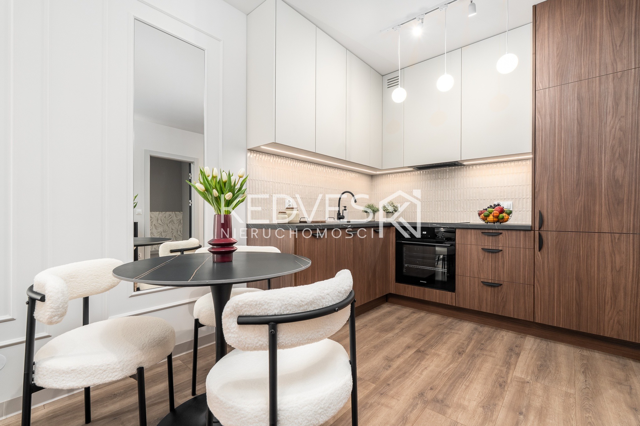 Nowoczesna kuchnia z drewnianymi i białymi szafkami, okrągłym stołem z kwiatami i pluszowymi krzesłami. Lustro odbija wnętrze, a oświetlenie podkreśla detale.