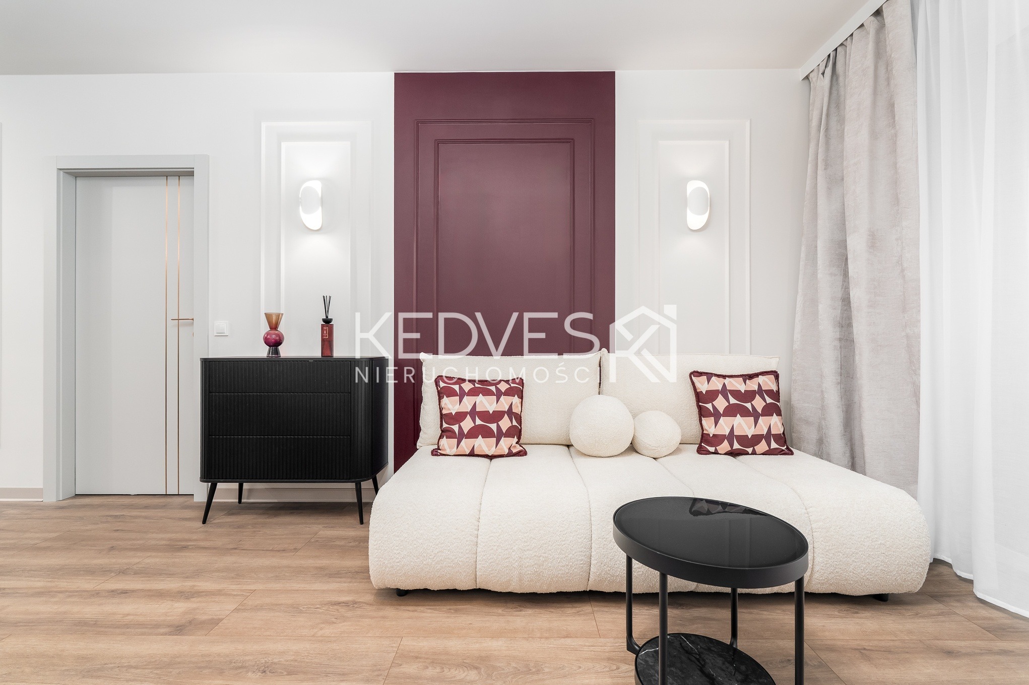 Stylowe wnętrze: kremowa sofa boucle, czarna komoda z dekoracjami, burgundowa ściana z panelami, drewniana podłoga i designerskie oświetlenie. Nowoczesny minimalizm.