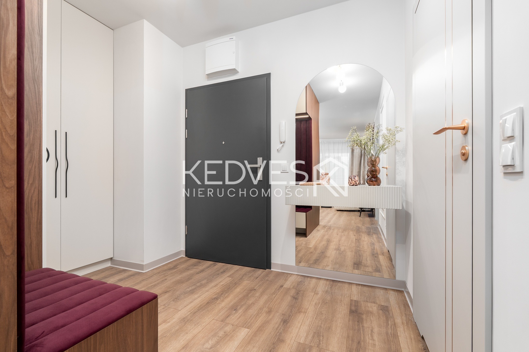 Nowoczesny przedpokój z szarą ścianą, lustrem w kształcie łuku, drewnianą podłogą i białą szafą. Widoczny fragment siedziska z bordową tapicerką.