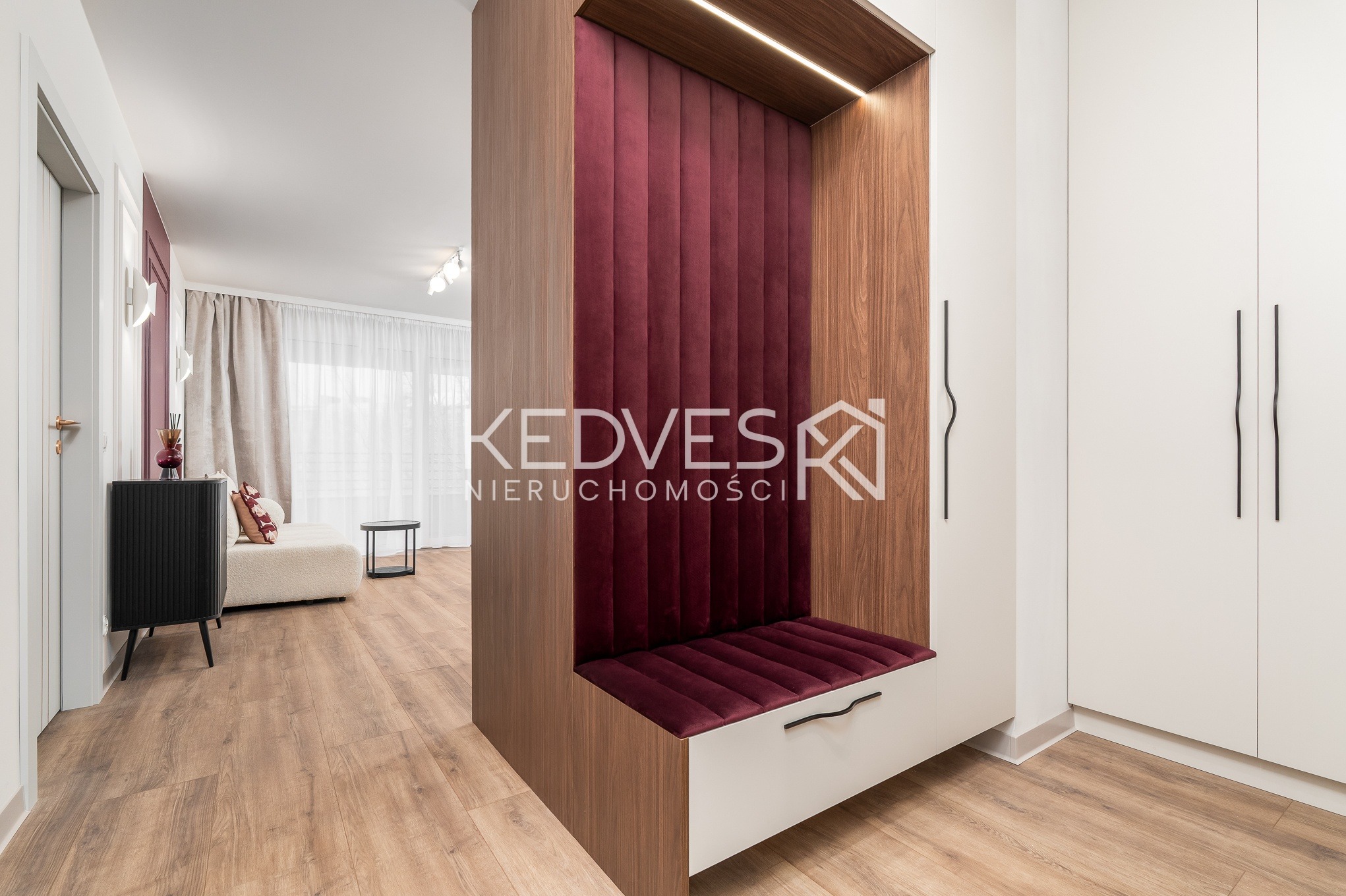 Nowoczesny przedpokój z siedziskiem w zabudowie z drewna i burgundową tapicerką. W tle widoczny salon z sofą i stolikiem. Całość utrzymana w jasnej kolorystyce.