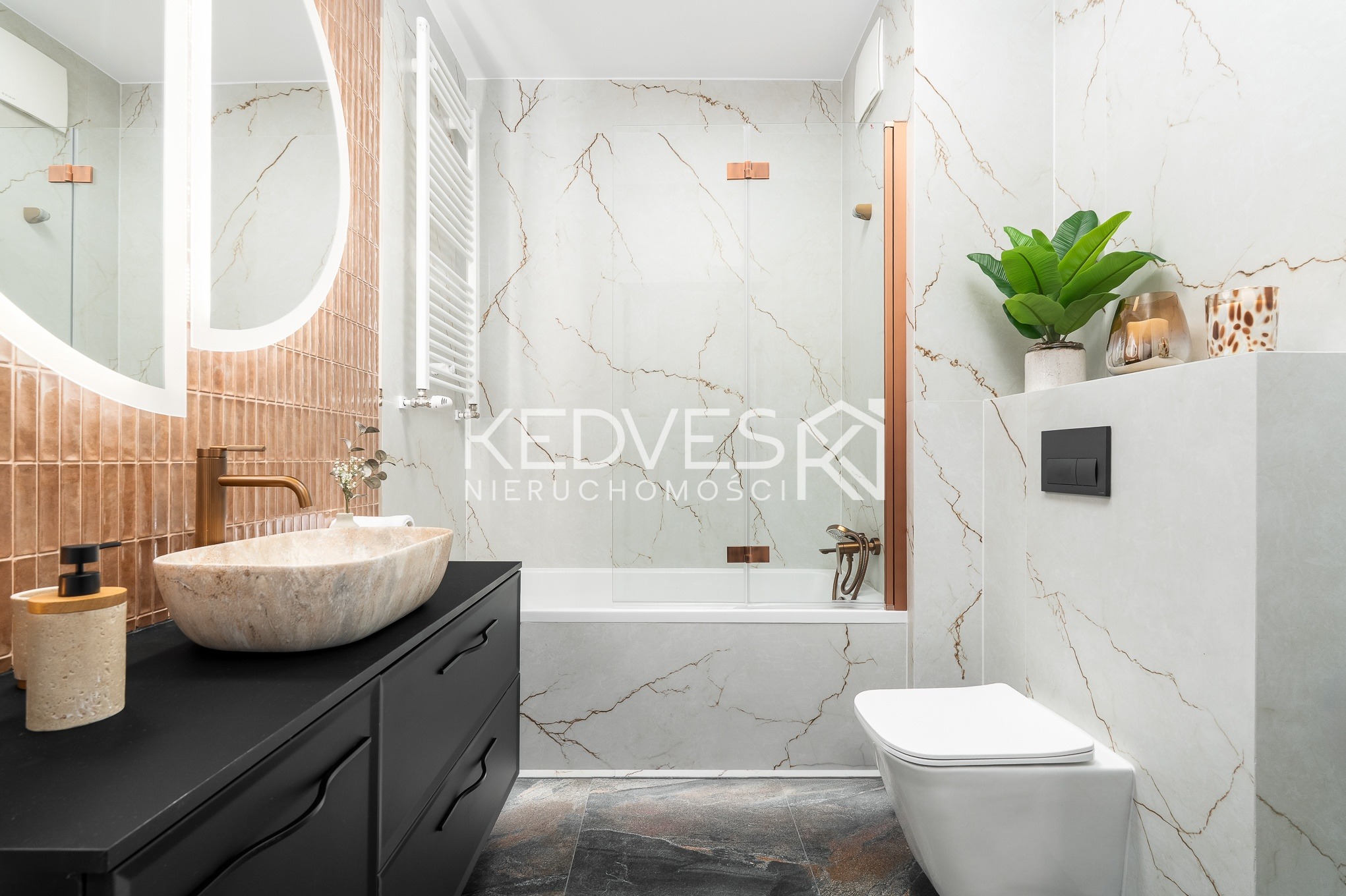 Elegancka łazienka z marmurowymi płytkami, owalną umywalką na czarnej szafce i złotą armaturą. Widoczna wanna za szklanym panelem i toaleta.