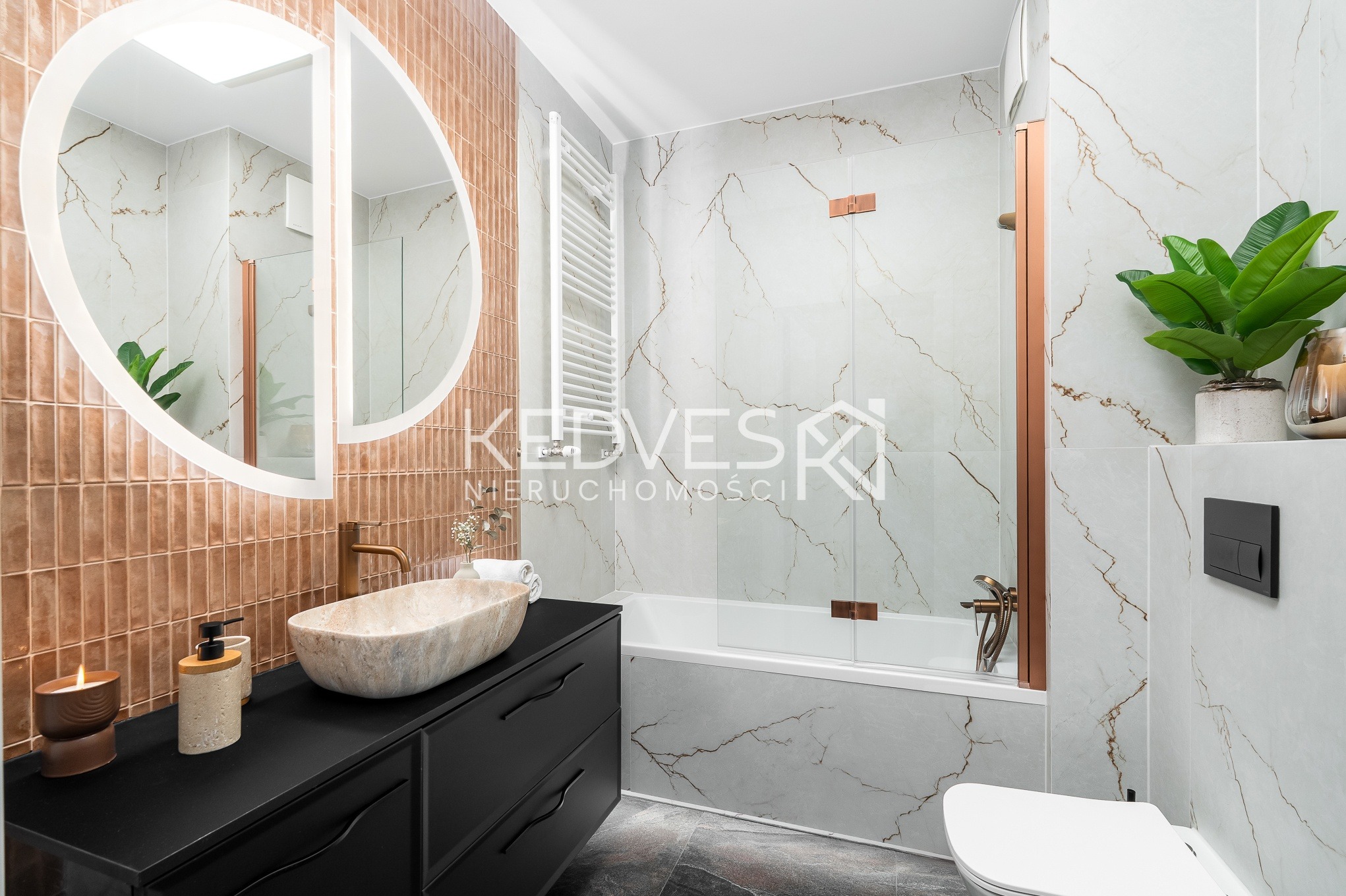 Nowoczesna łazienka z umywalką z kamienia, czarną szafką i okrągłym lustrem z podświetleniem. Beżowe płytki na ścianie i marmurowe płytki w strefie kąpieli.