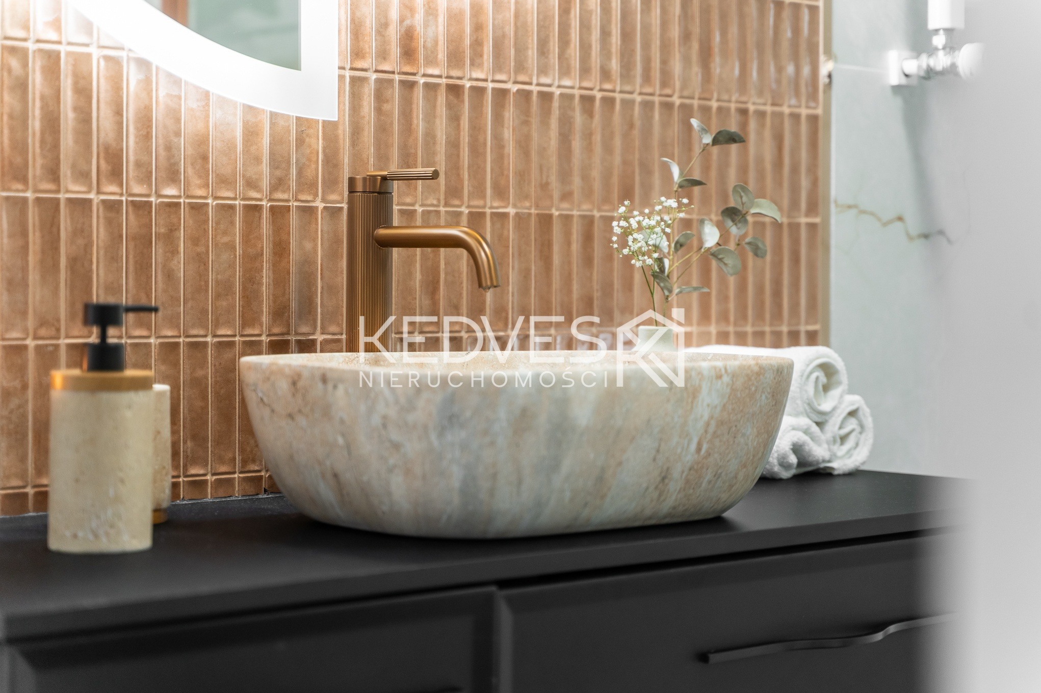 Stylowa łazienka: kamienna umywalka nablatowa, złota bateria, mozaika w odcieniach brązu i czarne szafki. Minimalistyczny design z akcentem natury.