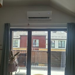 AmperCraft Tomasz Kendyk - Biała jednostka klimatyzacji Haier z wyświetlaczem 24, zamontowana na jasnej ścianie nad drzwiami balkonowymi. Wnętrze pokoju ze skośnym sufitem, widok na balkon i sąsiednie budynki.