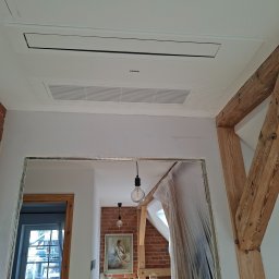 AmperCraft Tomasz Kendyk - Kasetowa jednostka klimatyzacyjna Haier w białym suficie, z kratką wentylacyjną. W tle wnętrze w trakcie prac, z odsłoniętymi drewnianymi belkami i ceglaną ścianą.