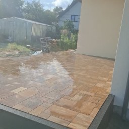 Profi-Bruk - Ułożona, mokra kostka brukowa w odcieniach brązu na tarasie domu jednorodzinnego, z widocznym obrzeżem i fragmentem elewacji w słoneczny dzień.