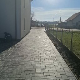 Profi-Bruk - Ułożona kostka brukowa w geometryczny wzór prowadząca wzdłuż budynku z szarą elewacją i ogrodzenia z siatki. Widoczny pracownik przy krawędzi.