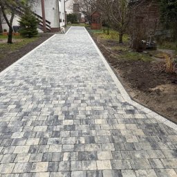 Profi-Bruk - Ułożona szara kostka brukowa prowadząca do domu z numerem 12, obrzeża z jasnego kamienia. Widoczny fragment ogrodu i drzewa bez liści. Pies na ścieżce.