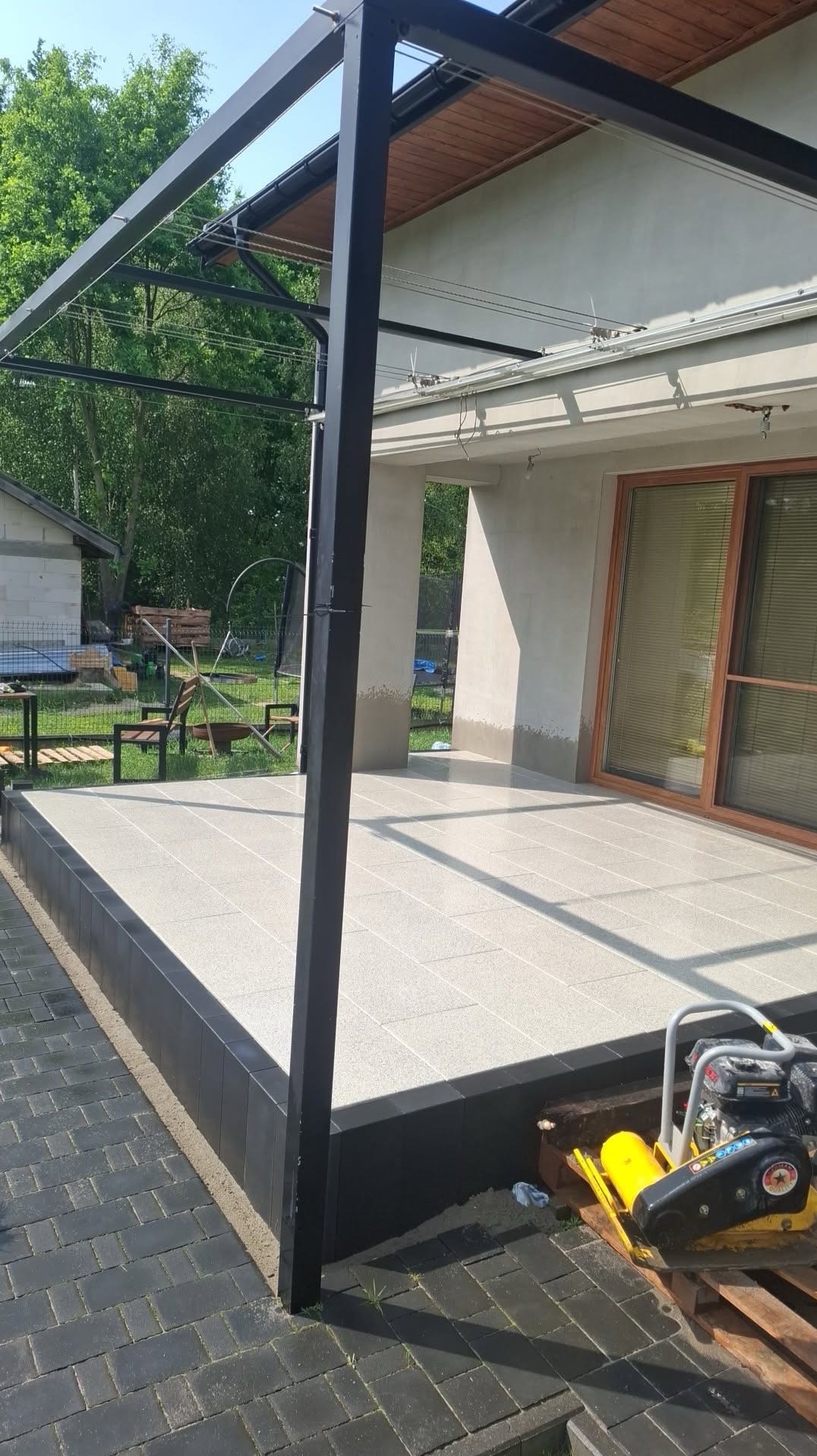 Jasna, wyłożona płytami tarasowa podłoga z czarną obwódką, z metalową konstrukcją zadaszenia. W tle dom z drewnianymi oknami i zielony ogród. Wibrator do ubijania kostki brukowej.