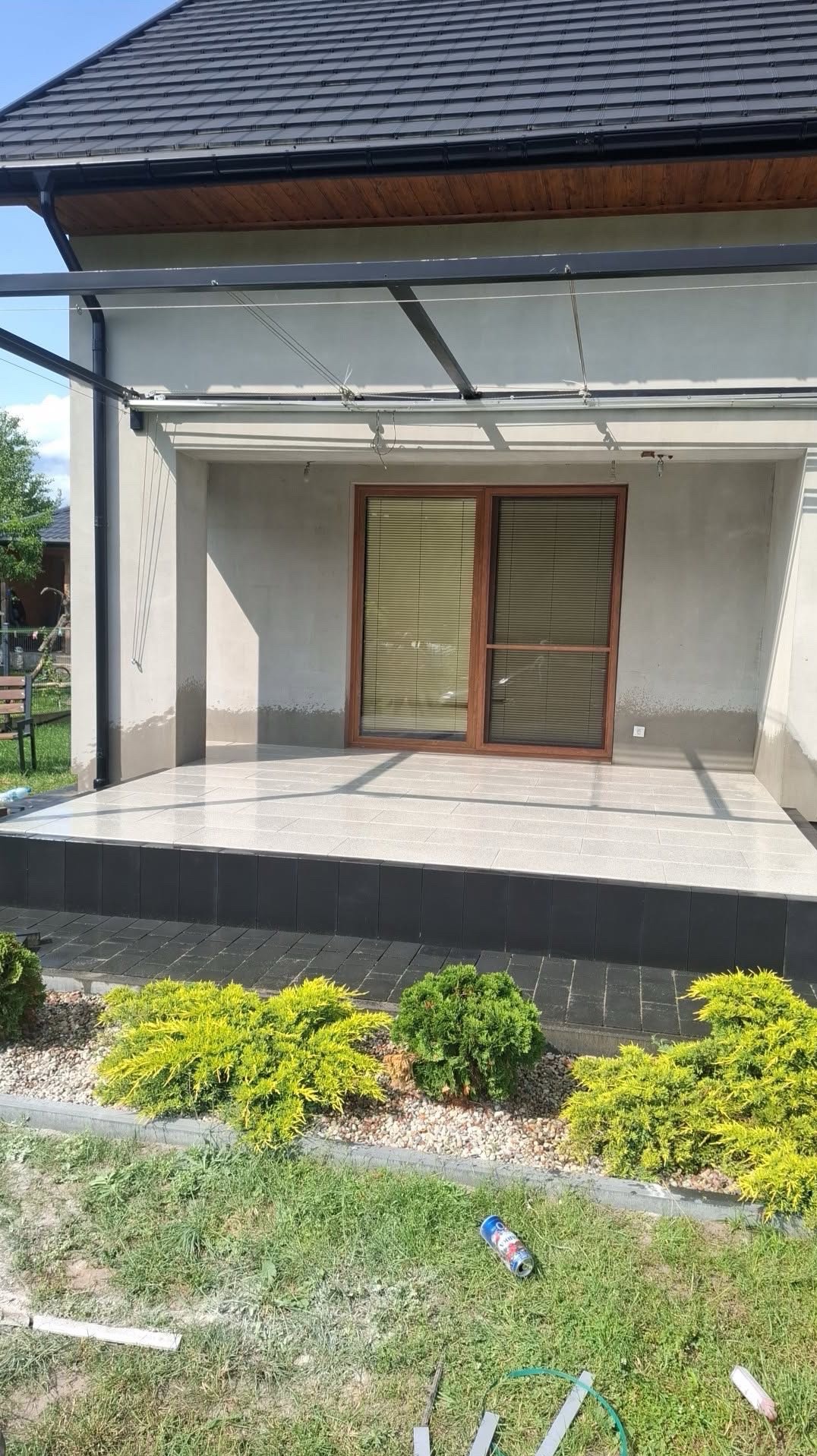 Zadaszony taras z jasną, kwadratową płytką, obrzeżem z ciemnej kostki i rabatą z krzewami. Widoczne elementy konstrukcji zadaszenia i fragment dachu domu.