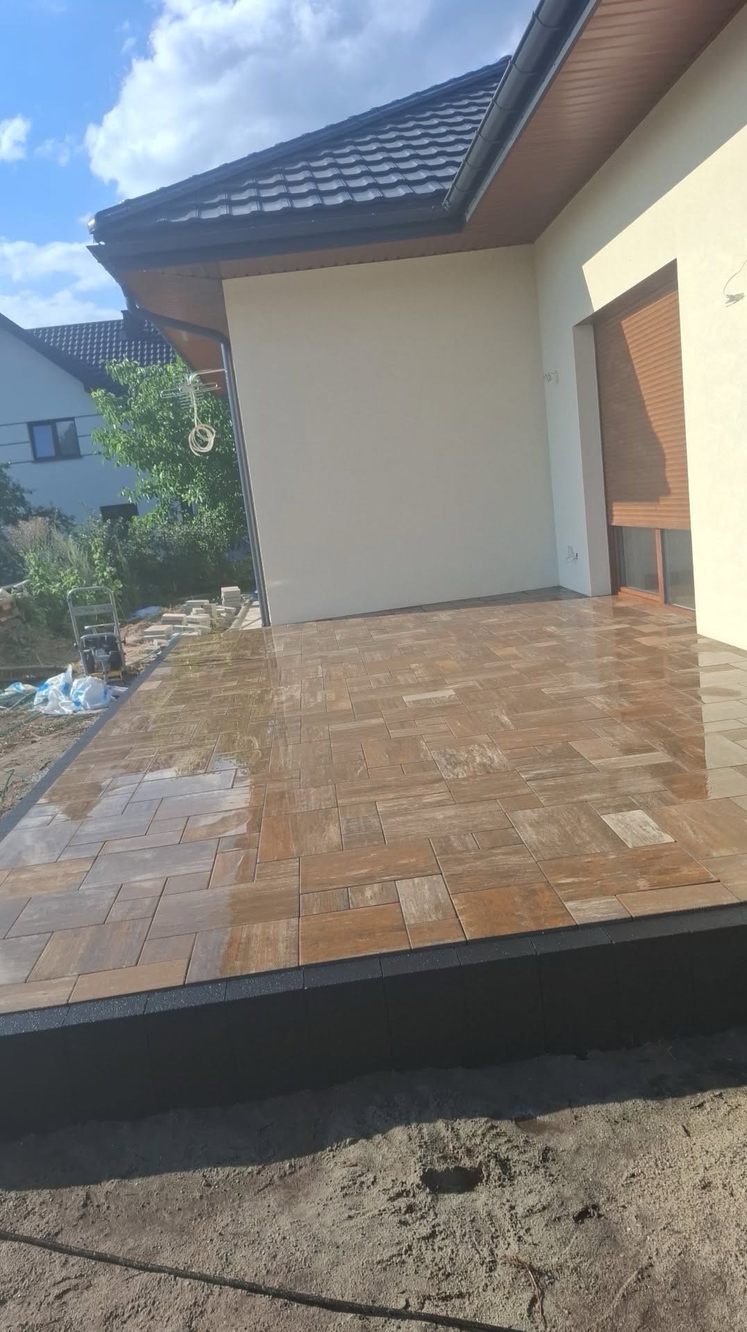 Ułożona, brązowa kostka brukowa na tarasie przydomowym, z widocznym obrzeżem i fragmentem elewacji budynku w tle. Częściowo mokra powierzchnia kostki.