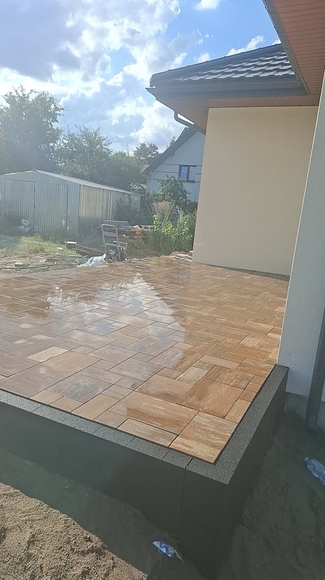 Ułożona, mokra kostka brukowa w odcieniach brązu na tarasie domu jednorodzinnego, z widocznym obrzeżem i fragmentem elewacji w słoneczny dzień.