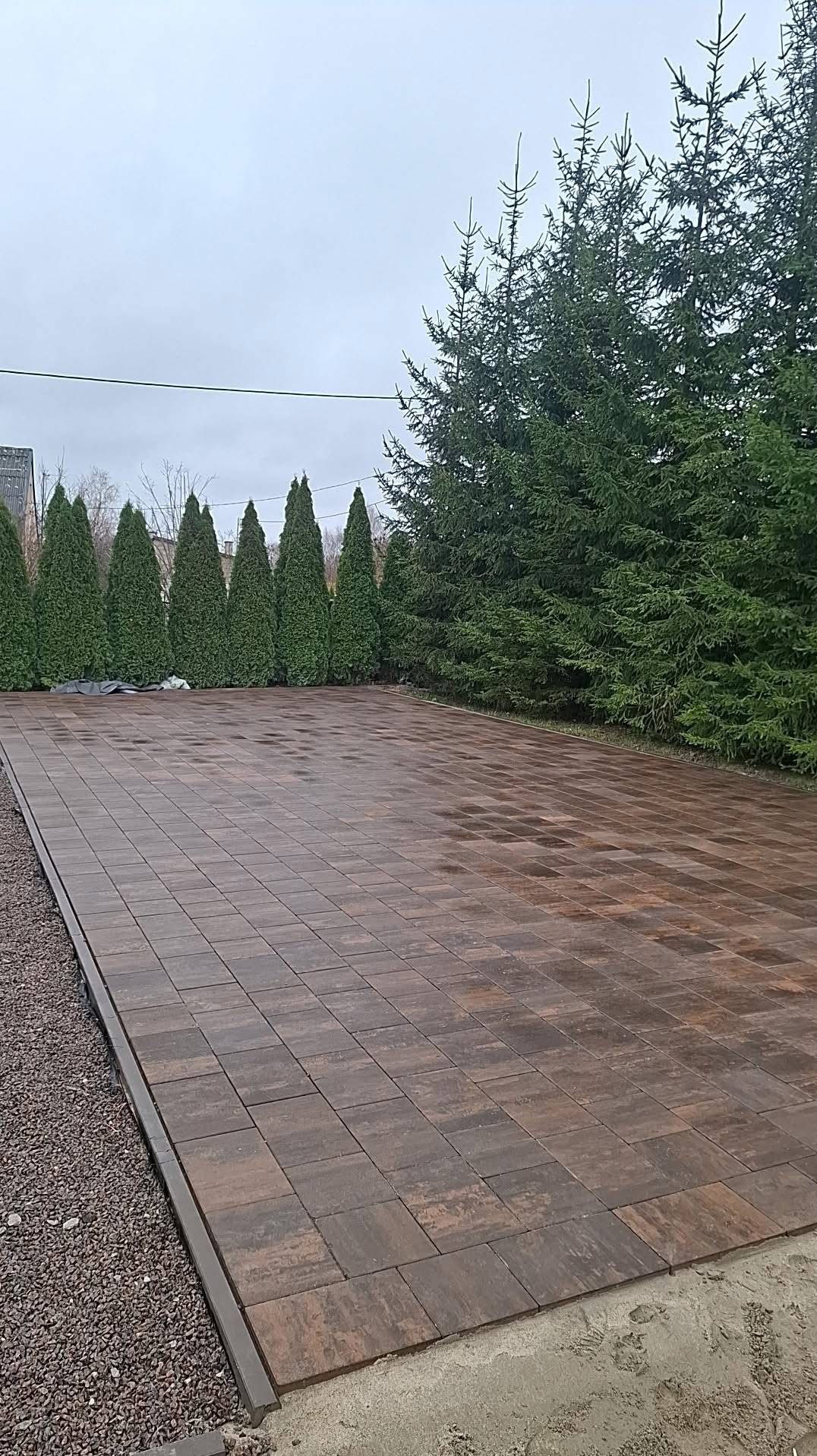 Ułożona, brązowa kostka brukowa na posesji z obrzeżem z grysu. W tle rząd tuj i wysokie drzewa iglaste. Pochmurne niebo.