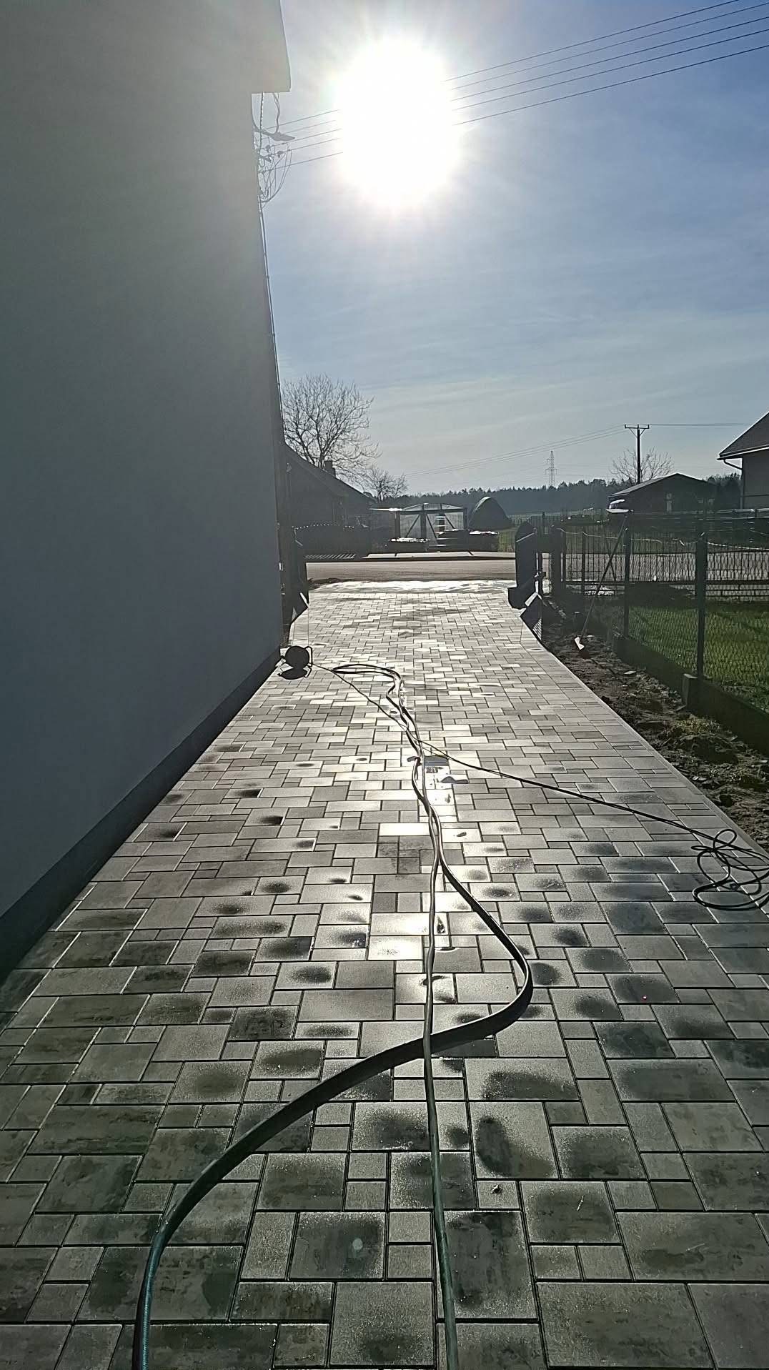 Świeżo ułożona, mokra kostka brukowa w odcieniach szarości, tworząca ścieżkę wzdłuż budynku. Widoczne kable i narzędzia, sugerujące trwające prace wykończeniowe.