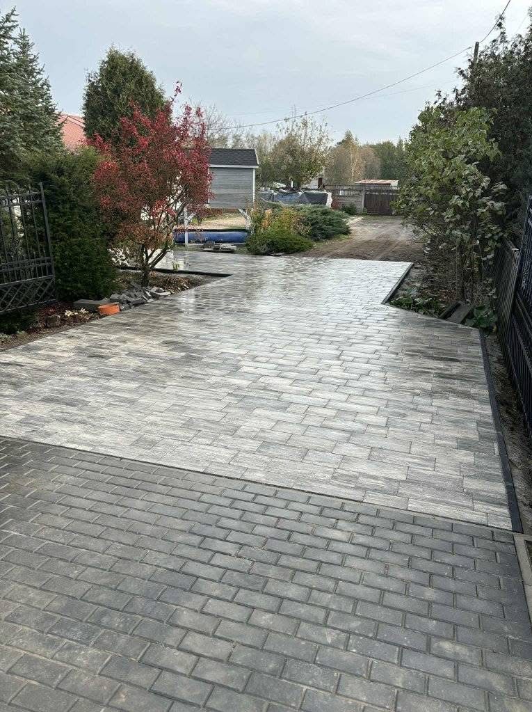 Ułożona, dwukolorowa kostka brukowa na podjeździe, z widocznym fragmentem ogrodu i ogrodzenia. Nowoczesny wzór kostki w odcieniach szarości i beżu.