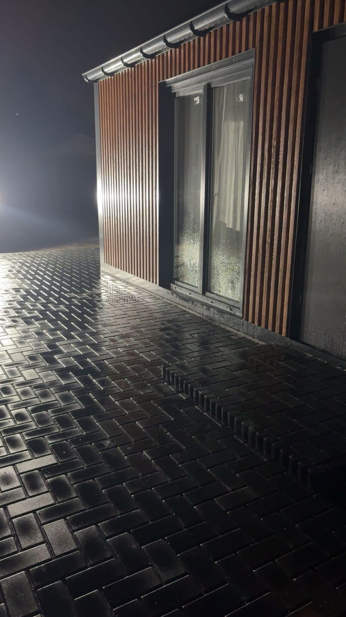 Ciemna kostka brukowa ułożona we wzór jodełki, mokra od deszczu, oświetlona nocą. Budynek z drewnianą elewacją w tle.