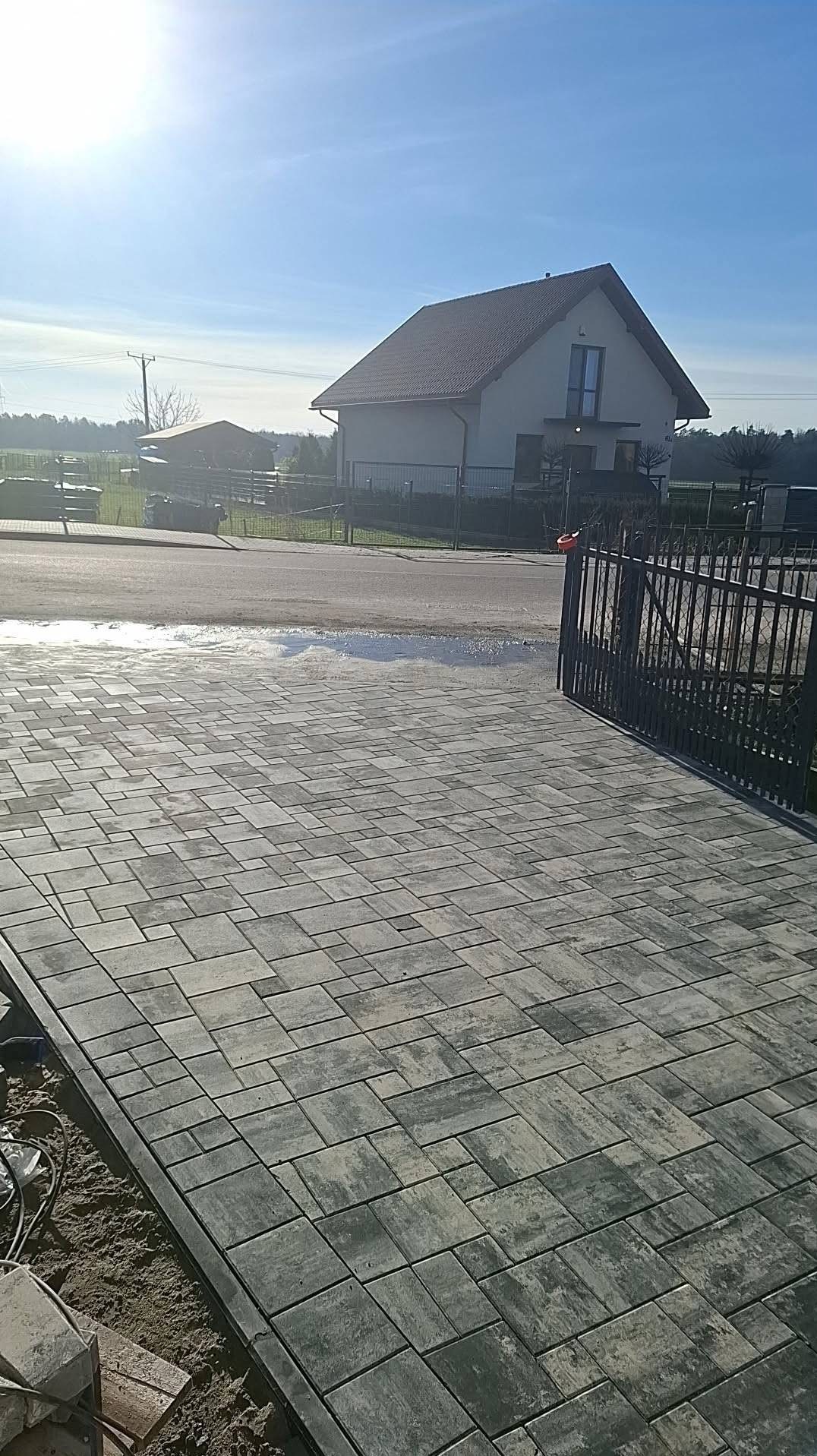 Ułożona kostka brukowa w odcieniach szarości prowadząca do bramy wjazdowej. W tle dom jednorodzinny i błękitne niebo.