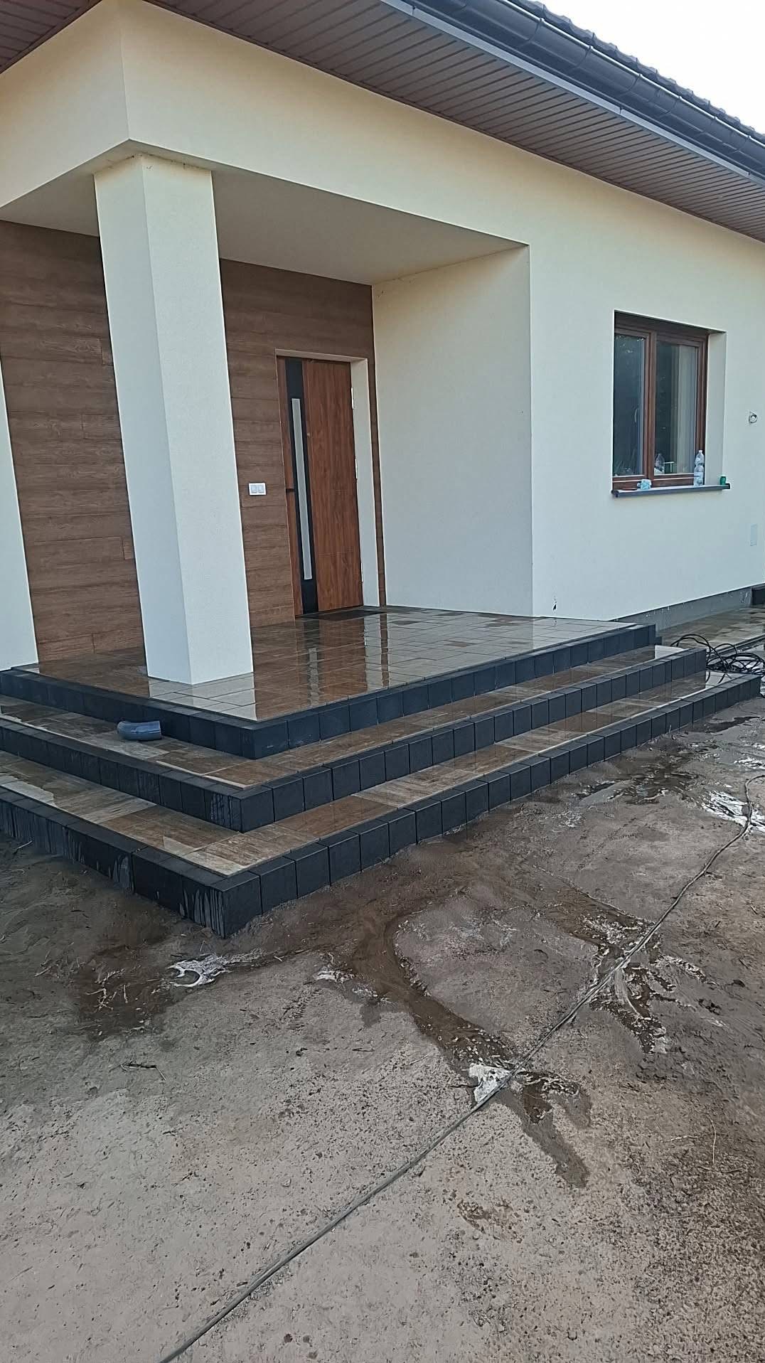 Wykończone schody wejściowe do domu z ciemną obwódką i jasnymi płytkami, mokre od wody. Widoczny fragment elewacji budynku i podłoże z resztkami błota.